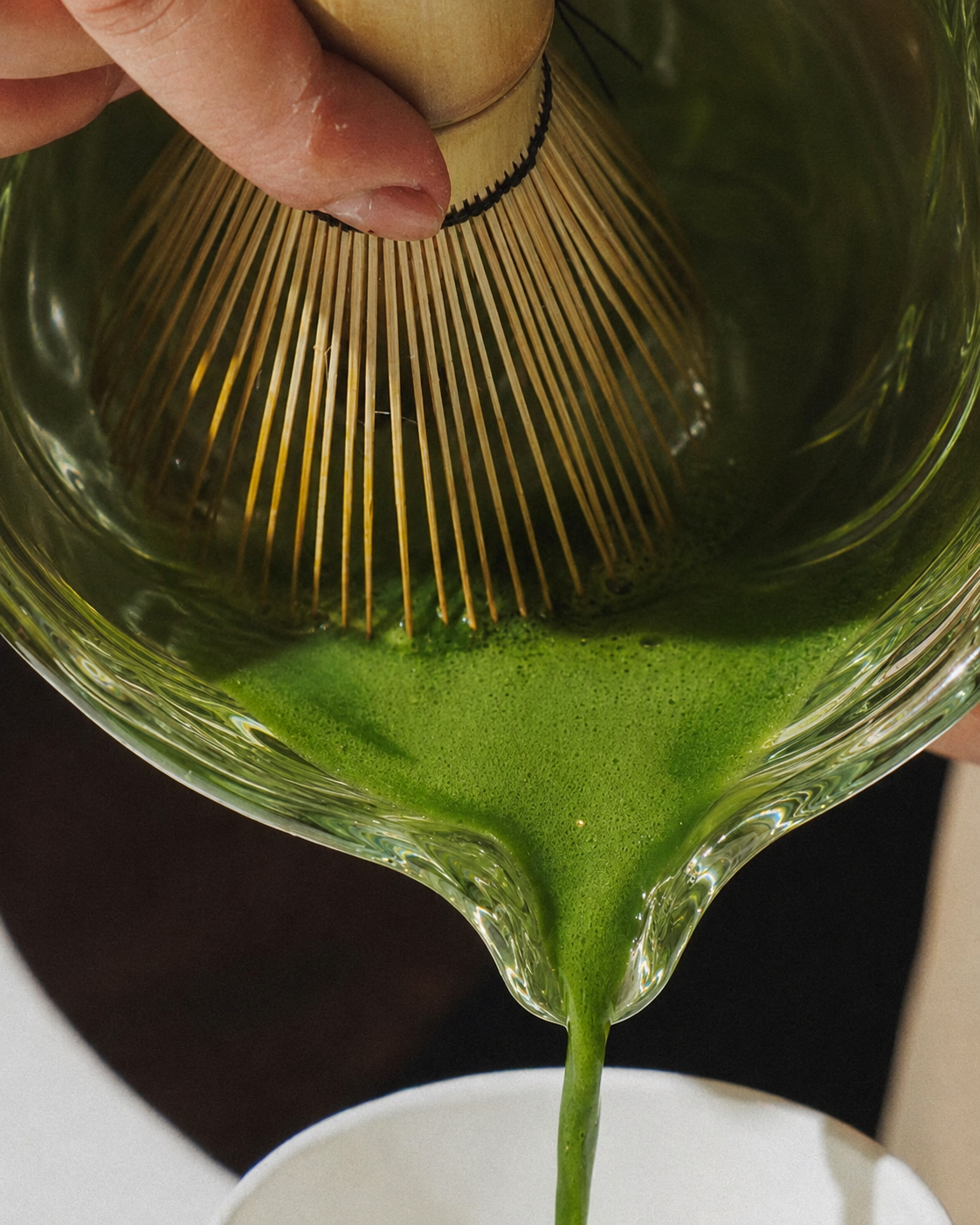 Vivid green matcha pouring from a ribbed glass bowl into a white cup.